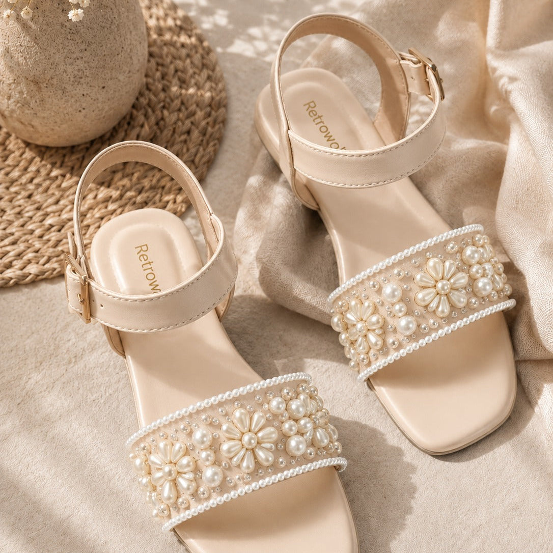 A pair of beige square-toe heeled sandals with ankle straps and wide embellished straps decorated with faux pearls and floral beadwork, styled on a beige fabric surface with a woven mat and dried flowers.