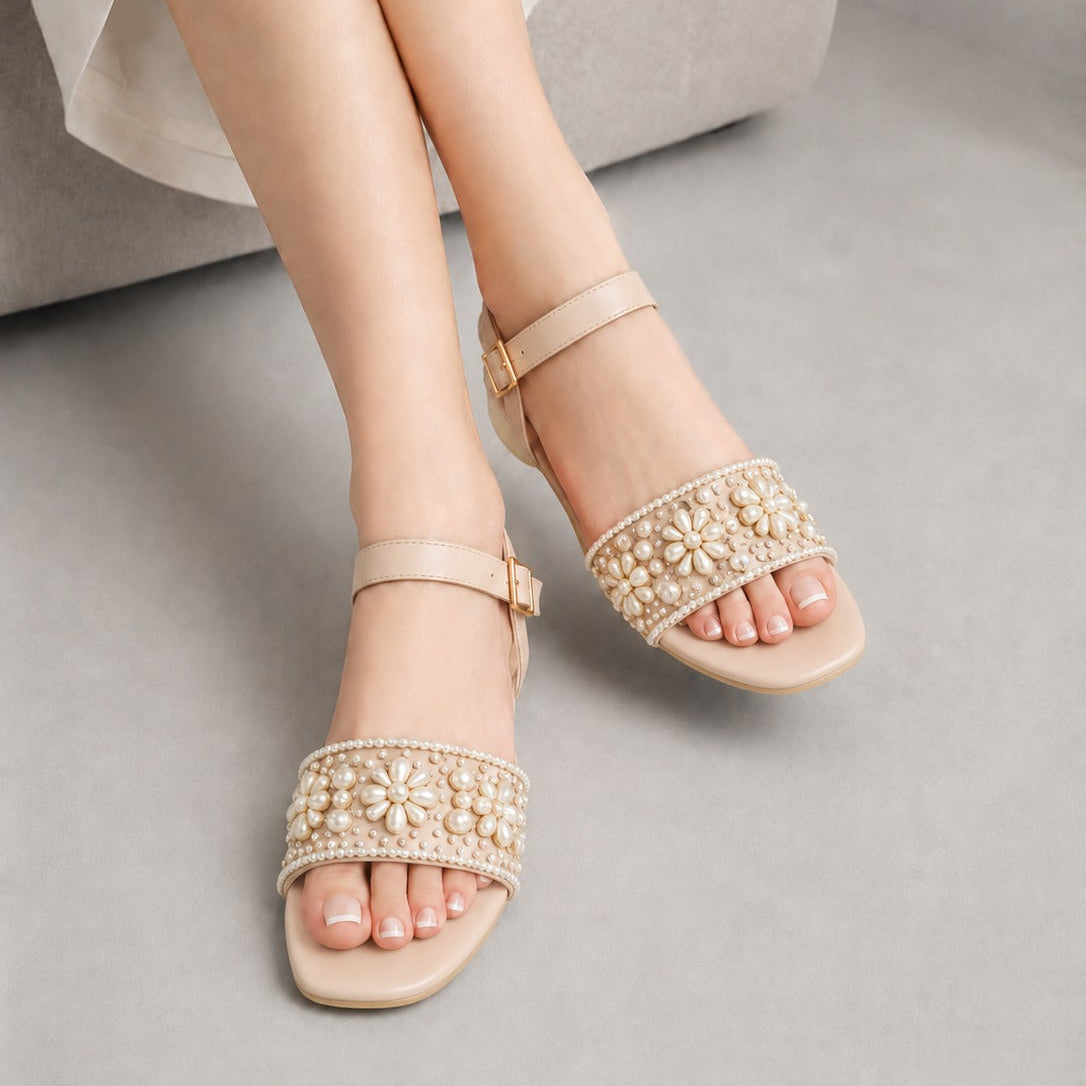 A person wearing beige flat sandals with wide embellished straps featuring pearl-like beads and rhinestone trim, seated on a light gray floor beside a gray upholstered chair.