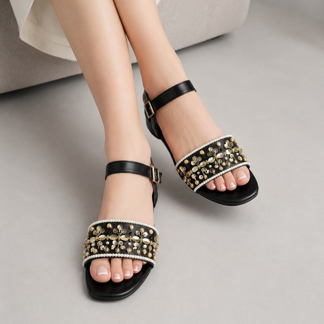 A person wearing black strappy flat sandals with wide embellished straps featuring gold studs, clear rhinestones, and white pearls, seated on a light gray sofa.