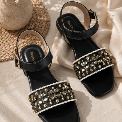 A pair of black flat sandals with ankle straps and wide embellished straps, styled on beige fabric with a woven mat and dried flowers. The sandals feature gold-tone and clear rhinestone embellishments with white pearl trim.