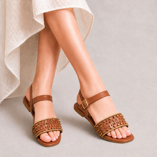 A pair of women's flat sandals with brown leather straps and woven tan accents on the toe straps, shown on a model wearing a light-colored dress against a neutral gray background.