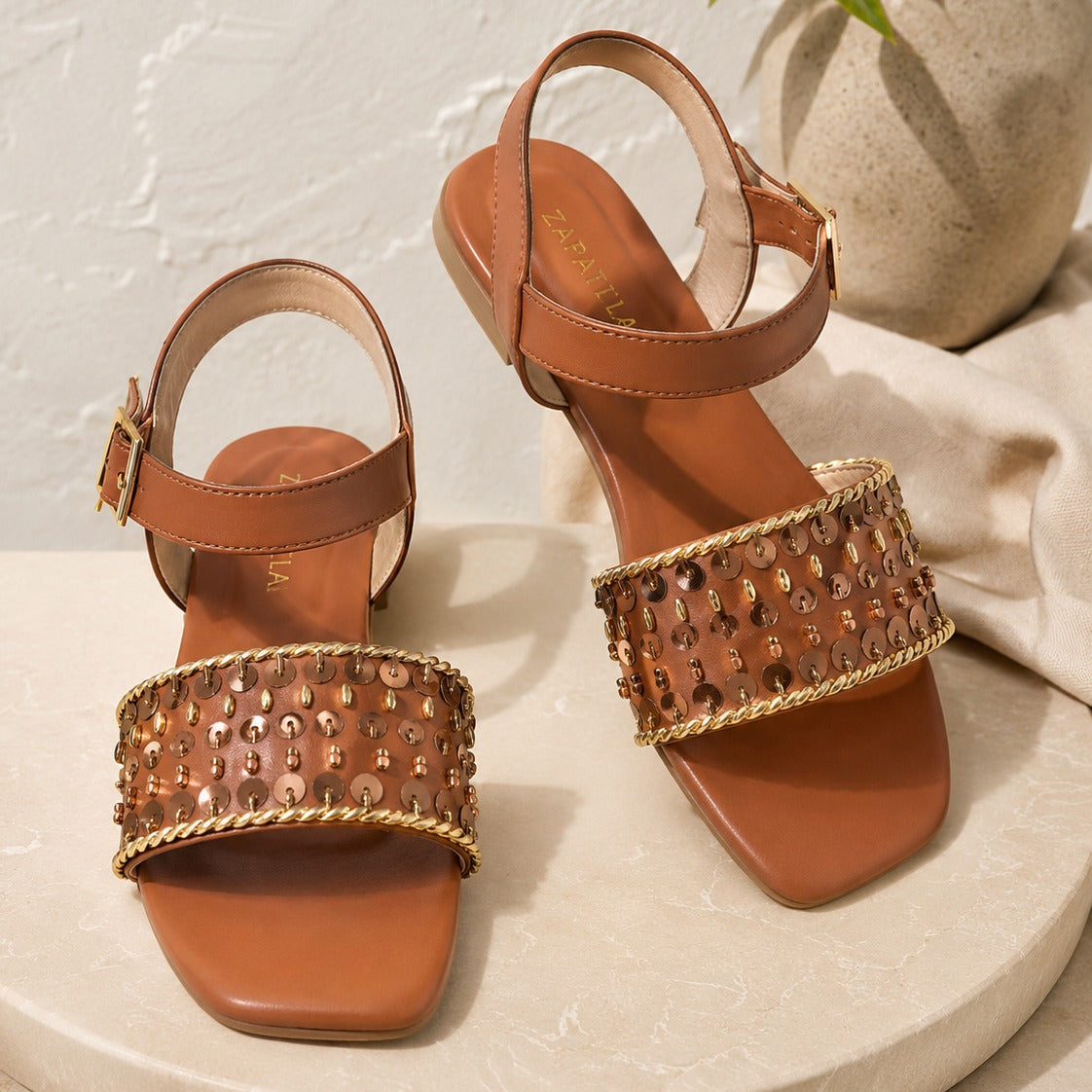 A pair of women's low sandals in tan leather, featuring a wide embellished strap with rhinestones and gold-tone chain trim, displayed on a round stone pedestal.